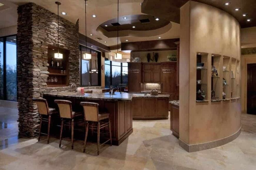 A stone brick pillar mounted with an inset shelf adds texture in this kitchen featuring granite countertops and wooden cabinetry that matches the island and peninsula. It is illuminated by cube pendants and recessed lights mounted on the tray ceiling.