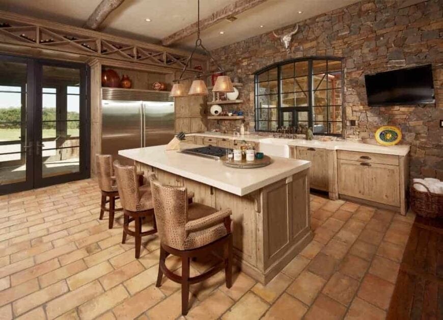 This kitchen boasts built-in wine racks arranged in a crisscross pattern along with wooden cabinets that match the breakfast island paired with wooden counter chairs. It has brick flooring and a stone accent wall mounted with a TV and an animal head decor.