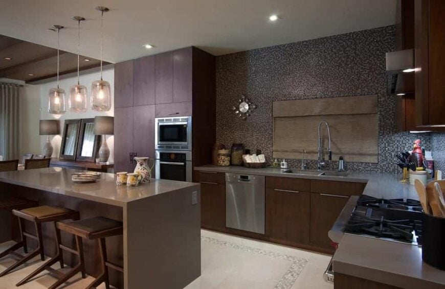 Mosaic tile backsplash mounted with a stylish clock adds a stunning accent in this contemporary kitchen with brown cabinetry and a sleek island lighted by glass pendant lights.