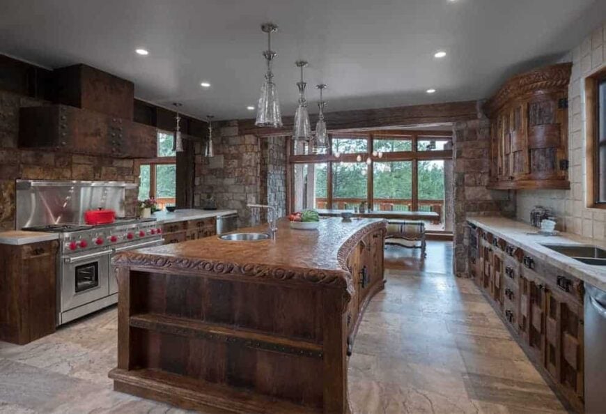 Rustic kitchen with stone brick walls and full height glazing allowing natural light in. It has stainless steel appliances and wooden cabinetry matching with the island that's illuminated by glass pendant lights.