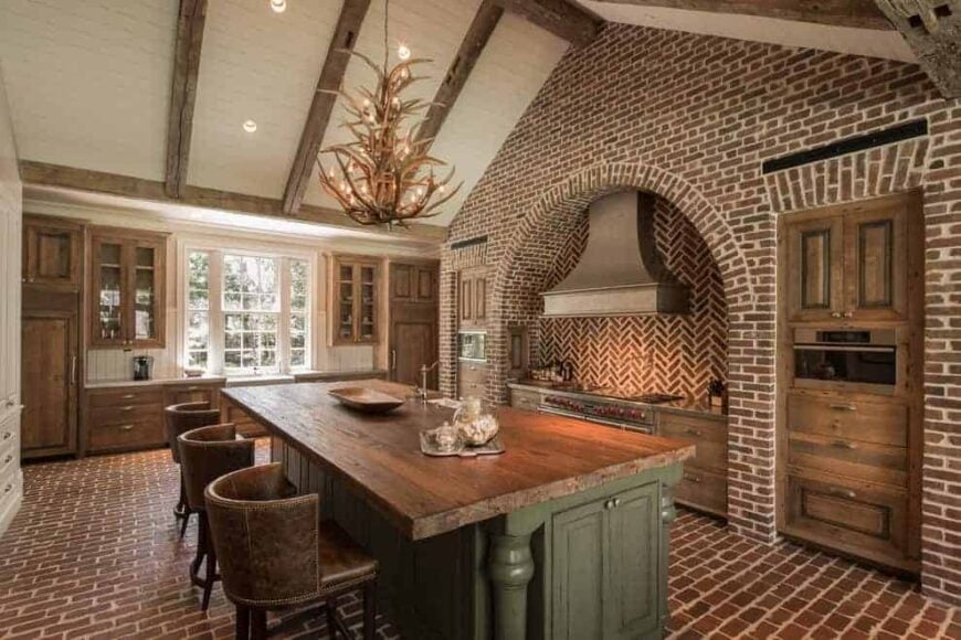 Round back counter chairs sit at a green island bar lighted by an antler chandelier that hung from the cathedral ceiling. It is accompanied by wooden cabinets and an arched cooking alcove with herringbone backsplash.