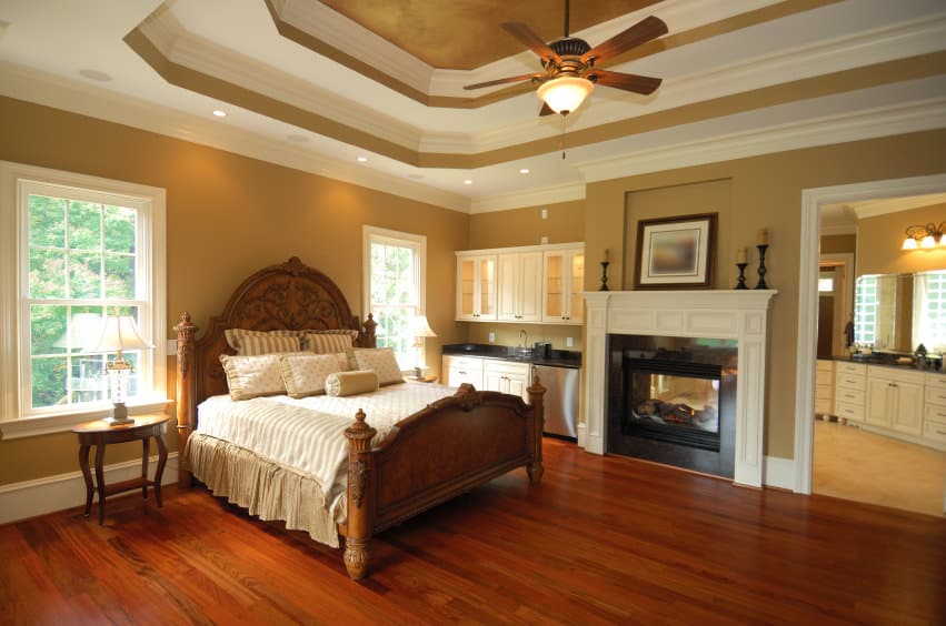 Primary bedroom featuring a stunning white and brown tray ceiling, along with brown walls and hardwood floors. The room offers a nice bed set and a fireplace on the side. There's a sink with cabinetry on the side. The room also has its own bathroom area.