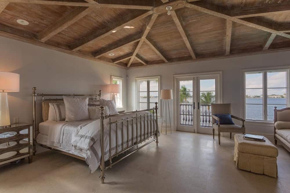 This is a charming and cozy primary bedroom with metal railings on its four-poster bed topped with a comfortable white cushion and pillows. There is also a peaceful sitting area at the other side of the room that is elevated by its consistent wooden ceiling and its exposed wooden beams.
