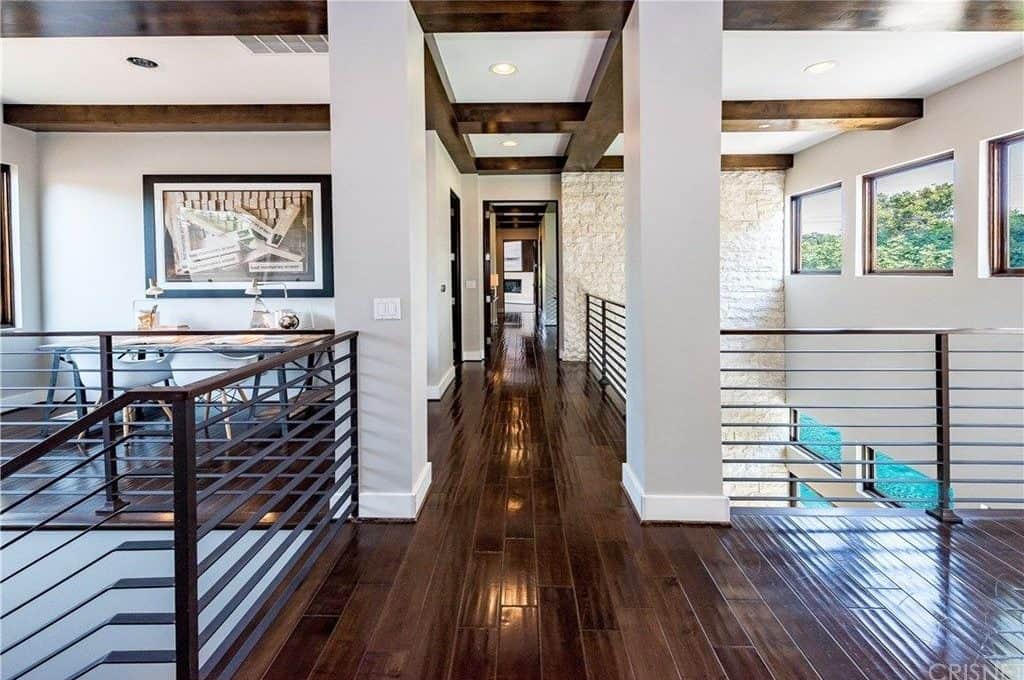 This is the second floor hallway of this home. It has a dark hardwood flooring that is contrasted by the flanking light gray columns that also contrast the dark wooden beams of the ceiling. This is augmented by the bordering dark iron railings.