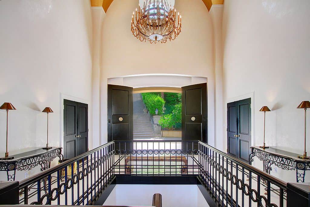 A black double door opens to this entry hall with a wooden bench and ornate console tables facing each other. It is illuminated by a classy candle chandelier that hung from the vaulted ceiling.