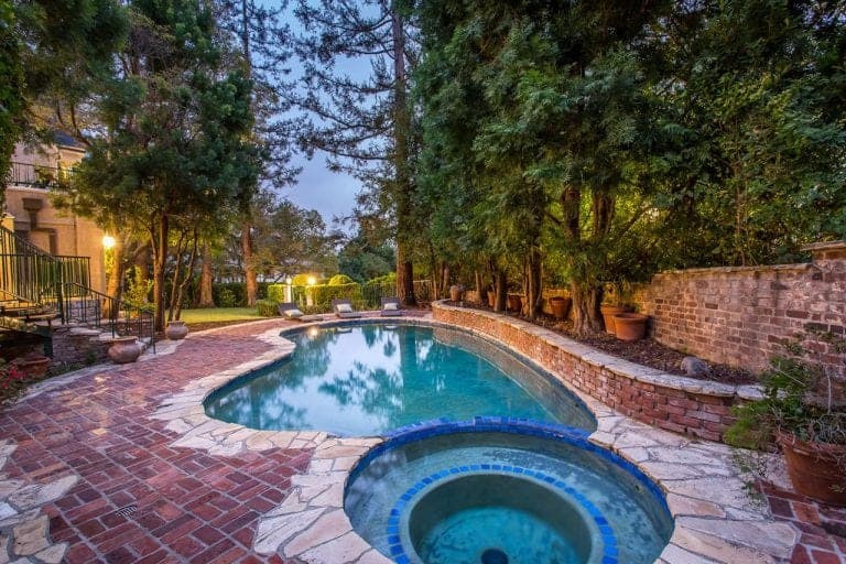 A gorgeous outdoor area featuring a stylish kidney-shaped swimming pool surrounded by tall mature trees. The outdoor space also has a garden area with well-maintained lawns.