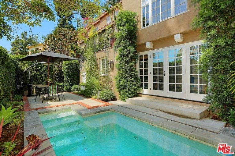This home offers a small swimming pool area with a table set on the side. The area is surrounded by green plants and trees.