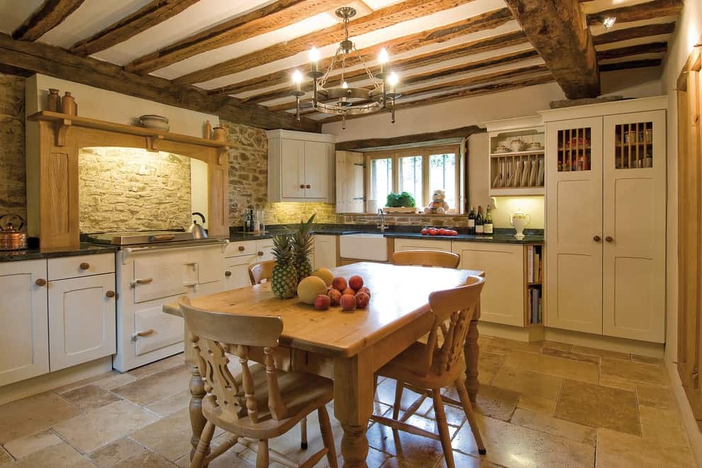 Country style kitchen offers white cabinetry and a cooking alcove against the brick backsplash. It has limestone flooring and wood beam ceiling mounted with a round chandelier.