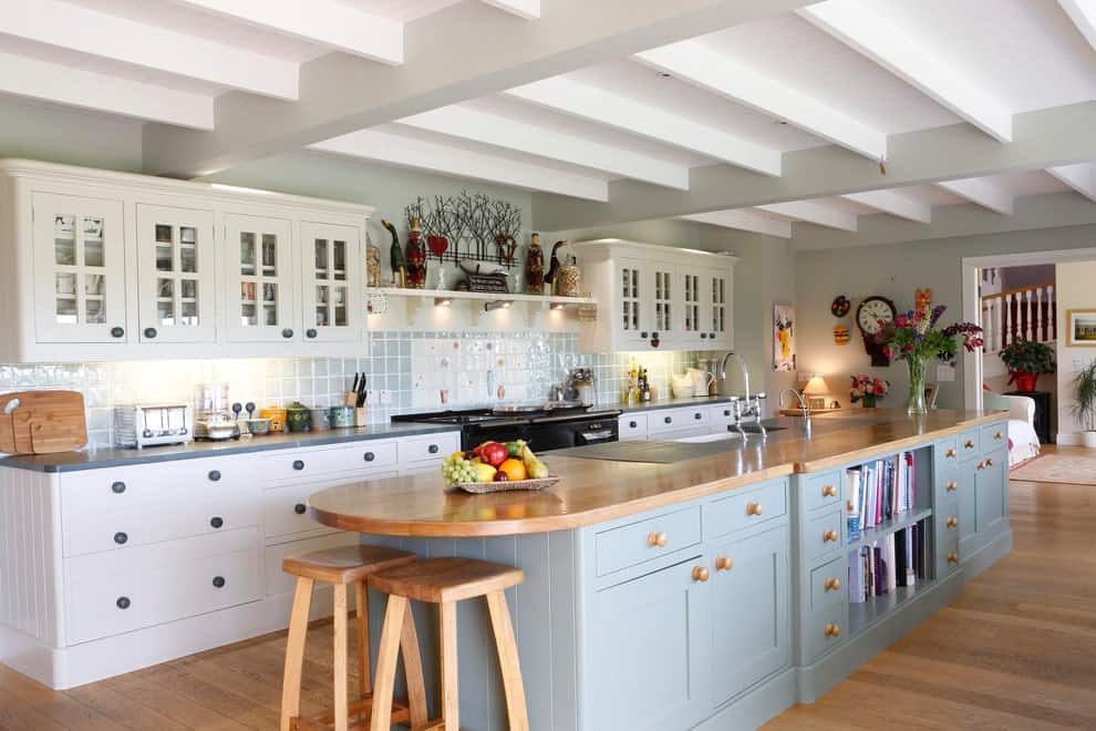 Cozy kitchen boasts white cabinetry and a grayish blue island bar complementing with the tiled backsplash. It is topped with a wooden counter that matches the bar stools and hardwood flooring.