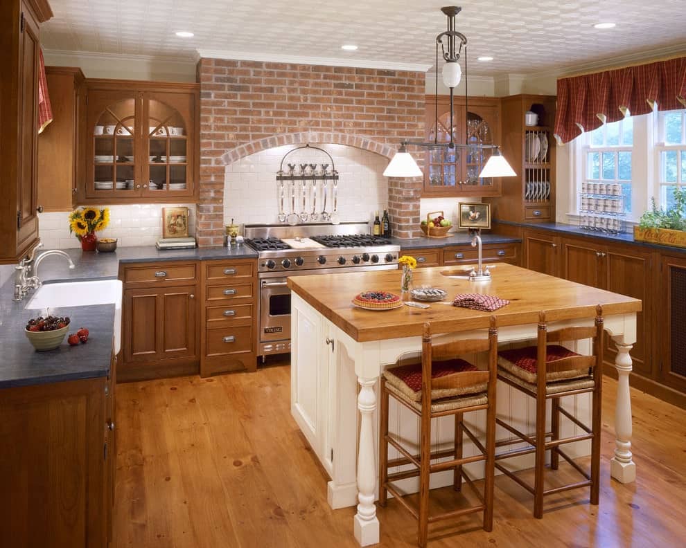 Glass dome pendants illuminate the white beadboard island that's paired with cushioned counter chairs. It is surrounded by a brick cooking alcove and wooden cabinetry that blends in with the hardwood flooring.