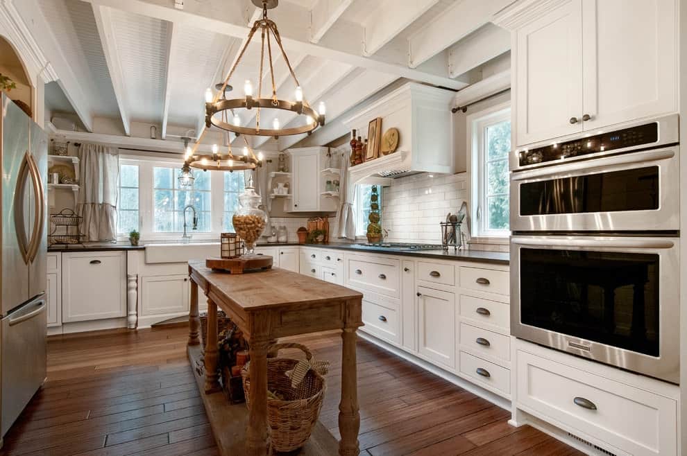 Stainless steel appliances and white cabinets surround a wooden island lighted by round chandeliers that hung from the beamed ceiling. This kitchen showcases wide plank flooring and white framed windows inviting natural light in.