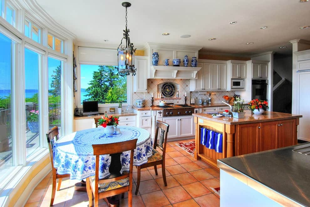 Country style kitchen offers white cabinetry and a wooden island bar along with a charming dining set illuminated by glass pendant light. It has terracotta flooring and glass paneled windows overlooking the outdoor scenery.