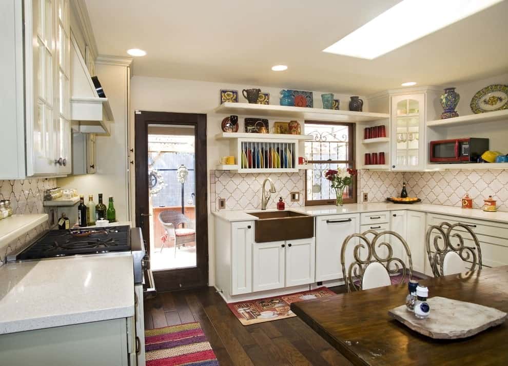 An eat-in kitchen with white cabinetry and floating shelves displaying gorgeous dinnerware. It includes a skylight and colorful rugs that lay on the wide plank flooring.
