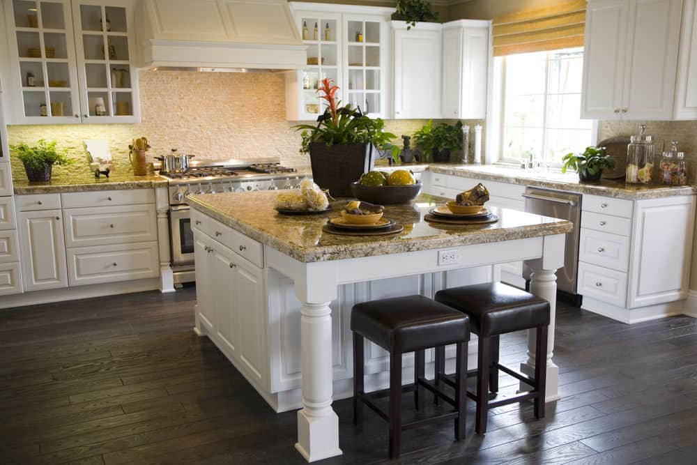 Stainless steel appliances and white cabinetry surround a granite top island paired with black cushioned stools. This kitchen has dark hardwood flooring and a glazed window covered in yellow roman shade.