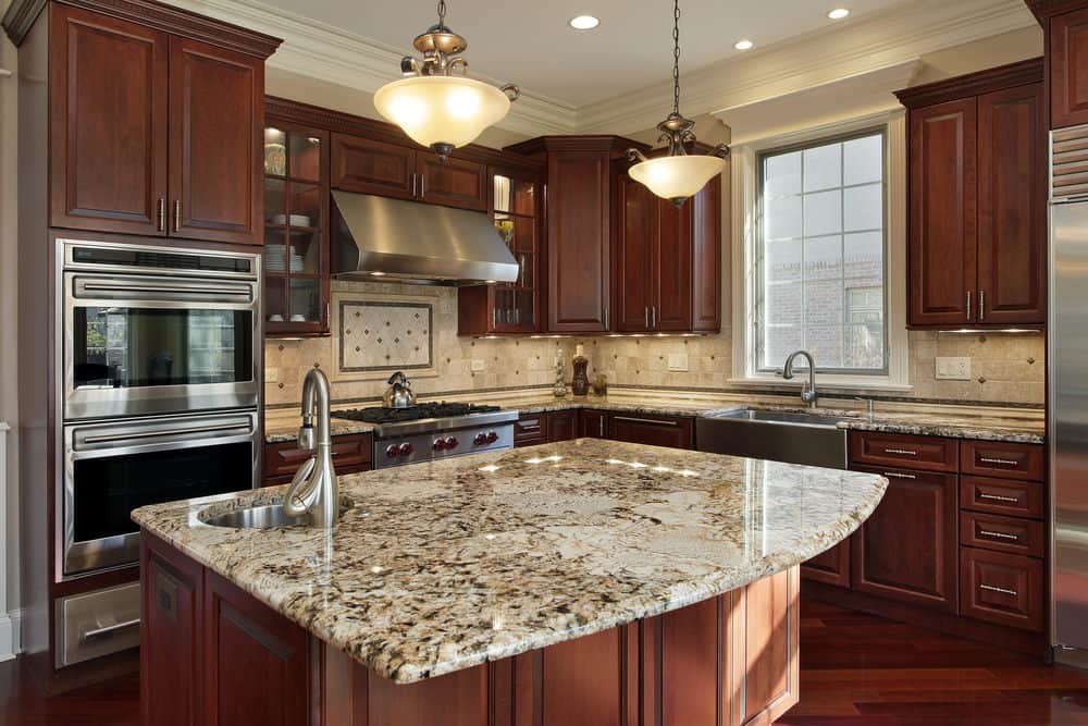 Glass dome pendants hang over the granite top island that's fitted with a round sink and chrome fixture. It complements the rich hardwood flooring and wooden cabinetry against the beige backsplash.