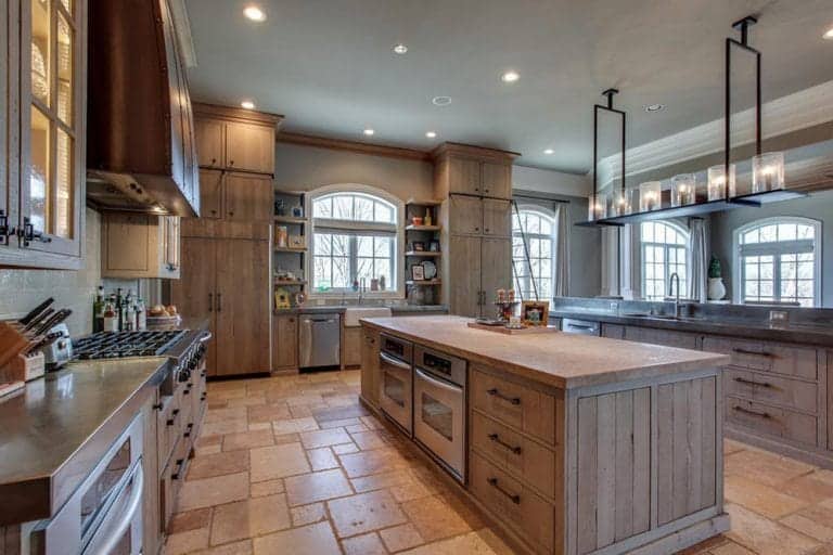 A large kitchen with light wood cabinets and two islands illuminated by a linear chandelier and recessed ceiling lights. It is surrounded by arched windows bringing natural light in.