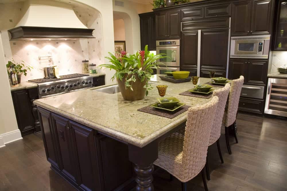 Dark wood cabinetry complements the breakfast island lined with wicker counter chairs. This kitchen offers stainless steel appliances and an arched cooking alcove completed with a dual fuel range and beige vent hood against the diamond pattern backsplash.
