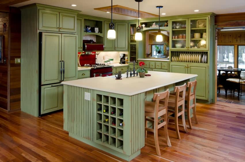 This kitchen boasts red appliances and green cabinetry matching with the quartz top island that's fitted with wine rack cubbies. It is lined with glass pendant lights and wooden counter chairs that blend in with the hardwood flooring.