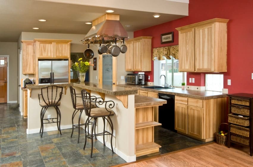 A stainless steel range hood integrated with metal pot rack stands over a two-tier kitchen island paired with ornate counter chairs. It is accompanied by inset appliances and light wood cabinetry against the white and red walls.