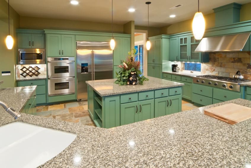 A curved peninsula along with green cabinetry and stainless steel appliances surround a granite top kitchen island that's fitted with built-in storage and shelves. It is illuminated by glass dome pendants and recessed ceiling lights.