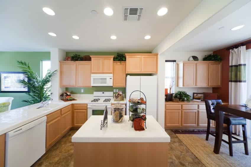 Eat-in kitchen with multicolored walls and tiled flooring topped by a wicker rug. It includes white appliances and wooden cabinetry matching with the kitchen island with a quartz countertop.