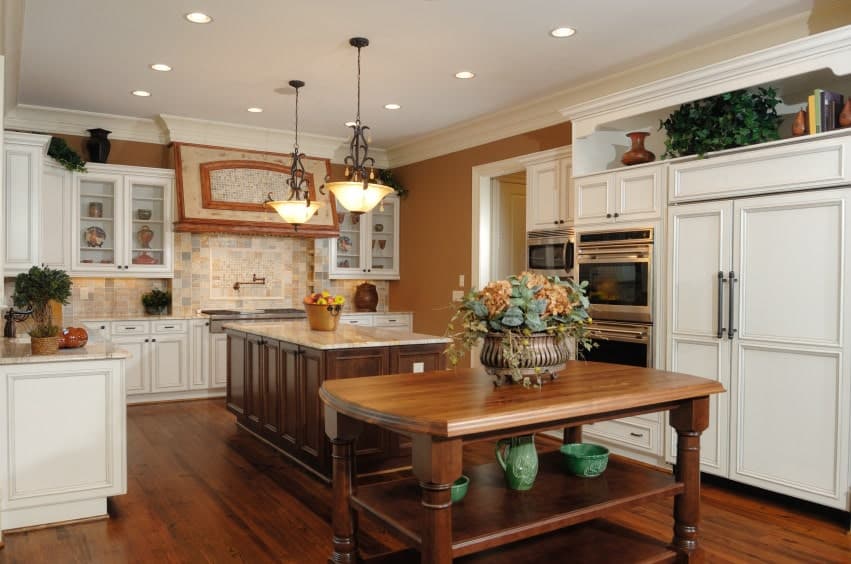A pair of wrought iron pendant lights illuminate the granite top island that blends in with the hardwood flooring. It is contrasted by white cabinetry against the brown walls.