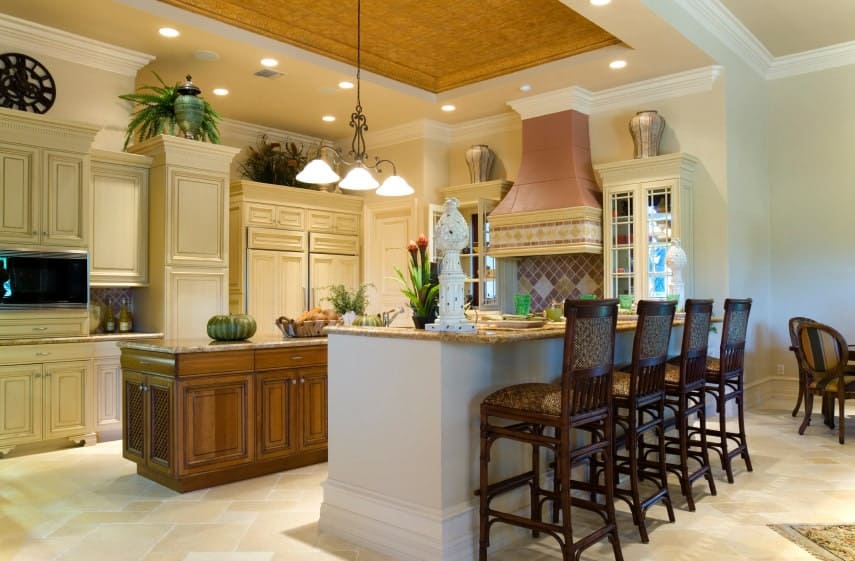 Printed cushioned chairs sit at a white breakfast bar that's topped with sculptures and granite counter. It is accompanied by beige cabinetry and a dark wood kitchen island lighted by glass dome pendants.