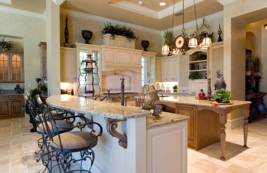 Country style kitchen features a two-tier breakfast bar and a wooden kitchen island over limestone flooring. It includes white cabinetry and a vintage chandelier that hung from the tray ceiling.