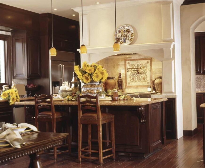 This kitchen is designed with a decorative plate and yellow pendant lights that hung over the dark wood island bar. It has stainless steel appliances and an arched cooking alcove against the brick backsplash.