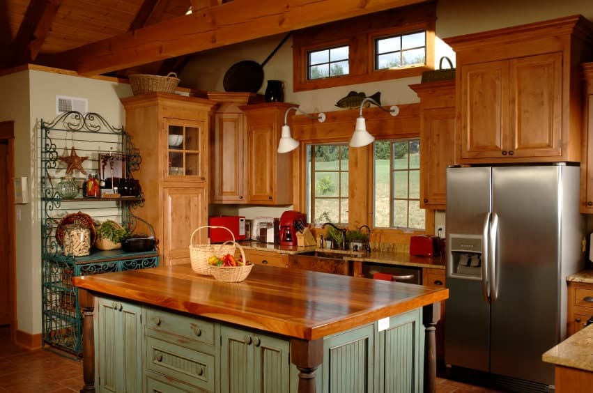 This kitchen showcases an ornate metal shelving and wooden cabinetry lighted by white sconces. There's a green breakfast island in the middle that's topped with a smooth wooden counter and rattan baskets. 