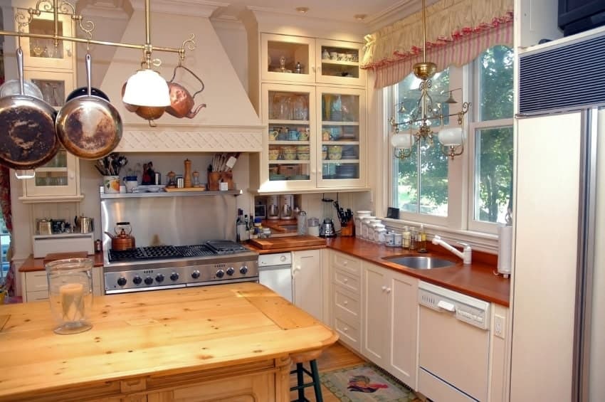 An ornate pendant light complements the brass pot rack that hung over the wooden kitchen island. This kitchen showcases white cabinetry and glazed windows dressed in a charming valance.