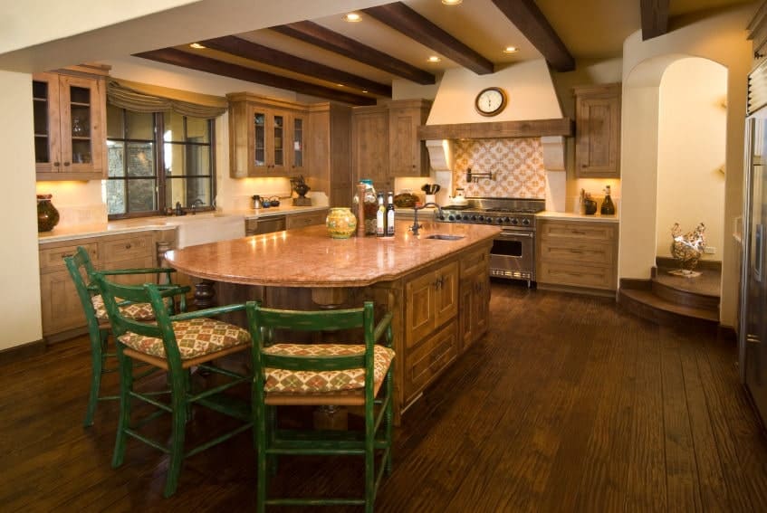 The cozy kitchen offers natural wood cabinetry and a granite top island paired with green cushioned chairs. It has wide plank flooring and beige ceiling lined with wood beams.