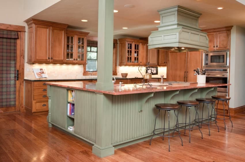 A green range hood matches the column and breakfast island with round bar stools. It is accompanied by stainless steel appliances and wooden cabinetry that blends in with the hardwood flooring.