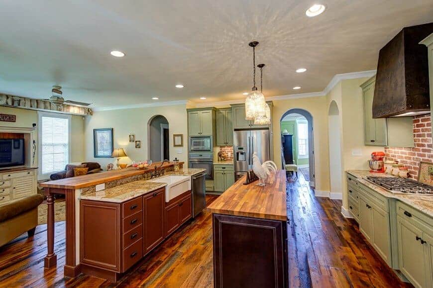 Glass dome pendants hang over the dark wood kitchen island that's situated in between a two-tier breakfast bar and green cabinetry. This kitchen features stainless steel appliances and a rustic vent hood fixed against the red brick backsplash.
