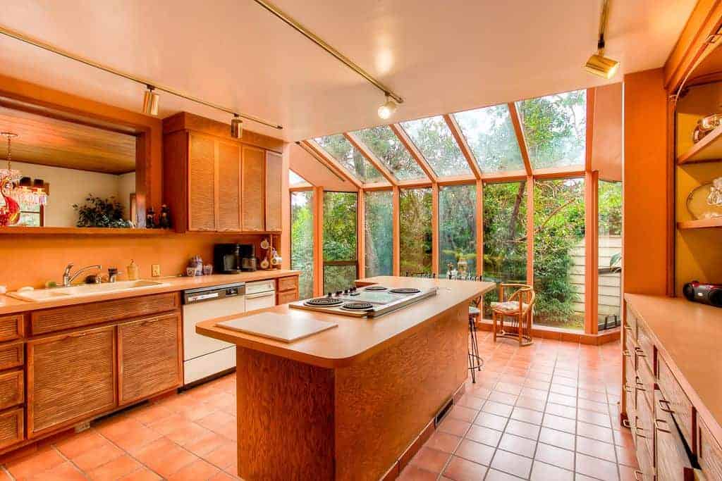 Warm kitchen with terracotta flooring and full height glazing overlooking the outdoor greenery. It includes white appliances and wooden cabinetry matching with the island bar that's fitted with a cooktop.