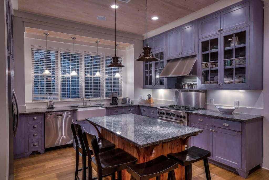 Black bar stools sit at a granite top island illuminated by dome pendant lights. It is surrounded by stainless steel appliances and purple cabinetry against the white walls.