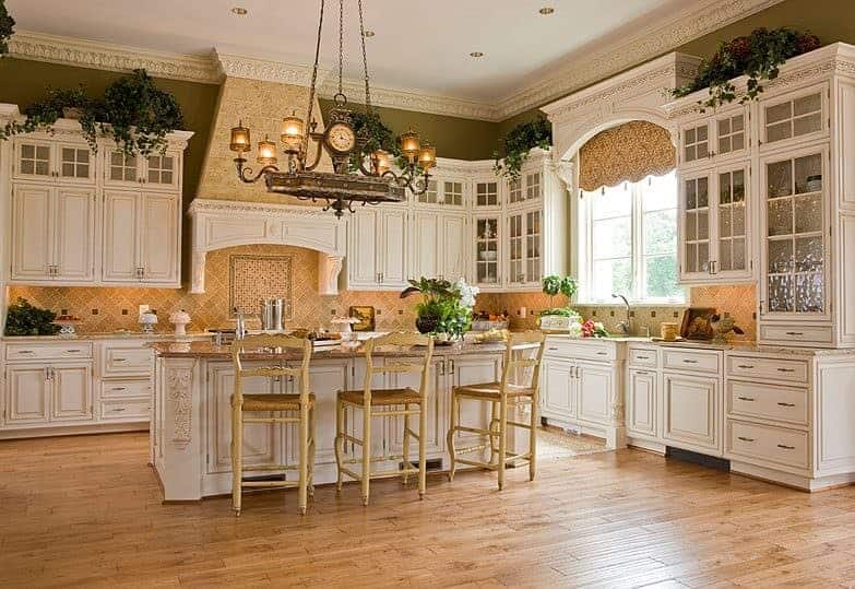 Sophisticated kitchen with white cabinetry and a matching island bar lighted by a vintage chandelier. It has light hardwood flooring and green walls lined with ornate crown molding.