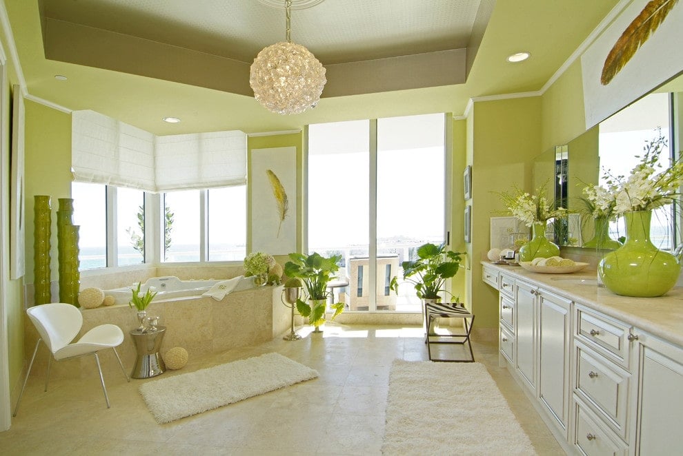 A contemporary white chair sits next to the corner tub facing the large vanity that's topped with apple green vases. It is illuminated by recessed lights and a round crystal pendant that hung from the tray ceiling.