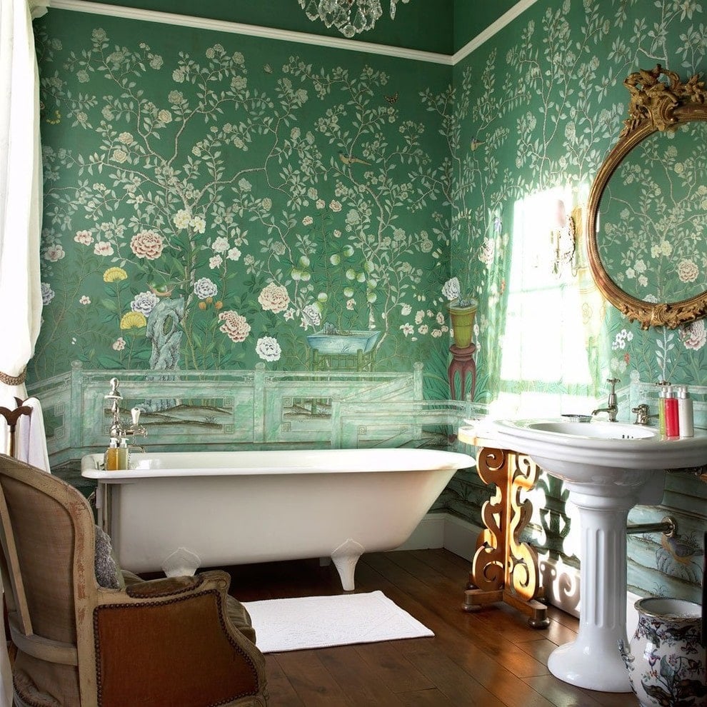 Clad in green floral wallpaper, this primary bathroom boasts a freestanding tub and a pedestal sink under an ornate round mirror. It is accompanied by a ceramic vase and a wooden armchair over wide plank flooring.