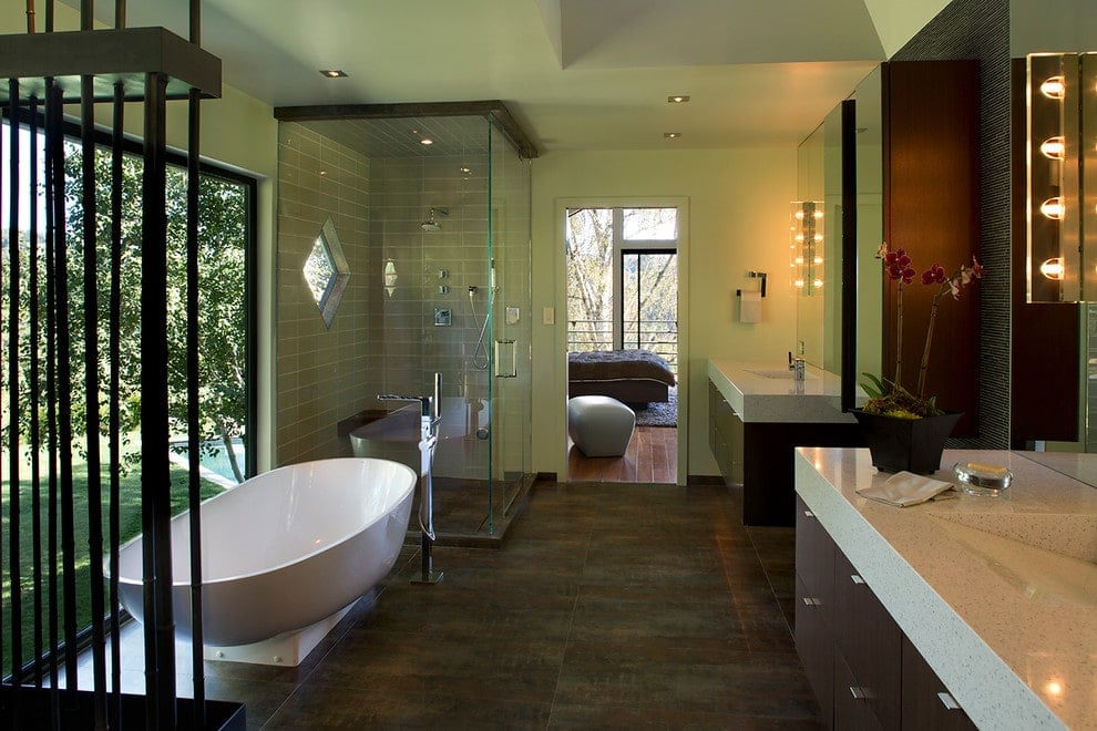 Linear glass sconces illuminate the floating sink vanities in this primary bathroom with distressed flooring and a panoramic window overlooking the lush green outdoor.