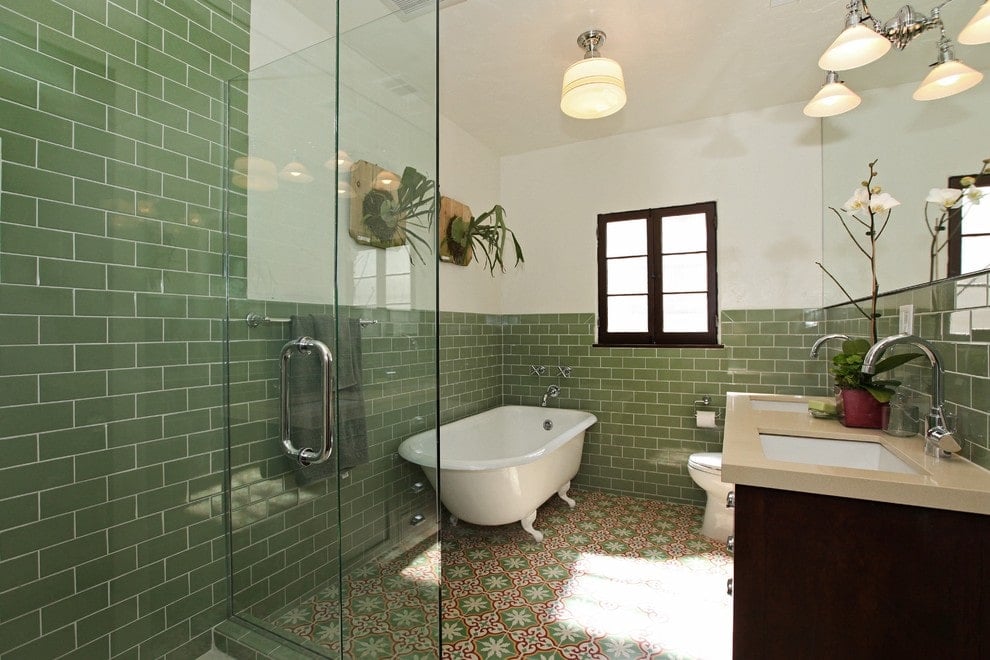 Fresh primary bathroom offers a clawfoot tub and a toilet that sits next to the dual sink vanity paired with a frameless mirror. It has decorative tile flooring and white walls dominated by green subway tiles.