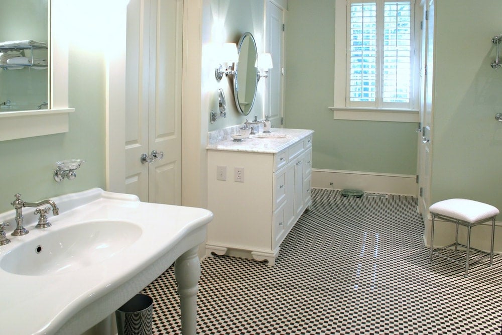 Black and white checkered flooring adds a striking contrast to the sage green walls in this primary bathroom with a porcelain washstand and white vanity under a round mirror flanked by chrome sconces.