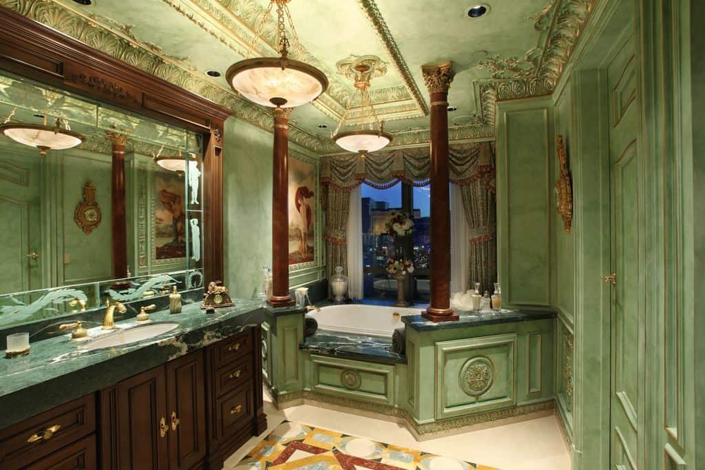 Deluxe primary bathroom with green wainscoted walls and an intricate tray ceiling mounted with brass pendant lights. It includes a large vanity and an alcove tub lined with brown columns.