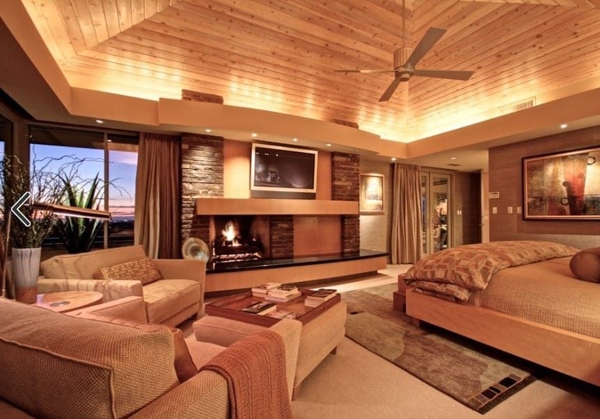 Large primary bedroom boasting brown walls, brown carpet floors and a brown wooden ceiling. The room offers a brown bed set and a fireplace, along with a sitting area on the side.