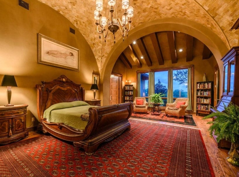 A spacious primary bedroom boasting a stunning bed set on top of a massive red area rug covering the hardwood flooring. The room has a stunning groin vault ceiling lighted by stylish chandelier.
