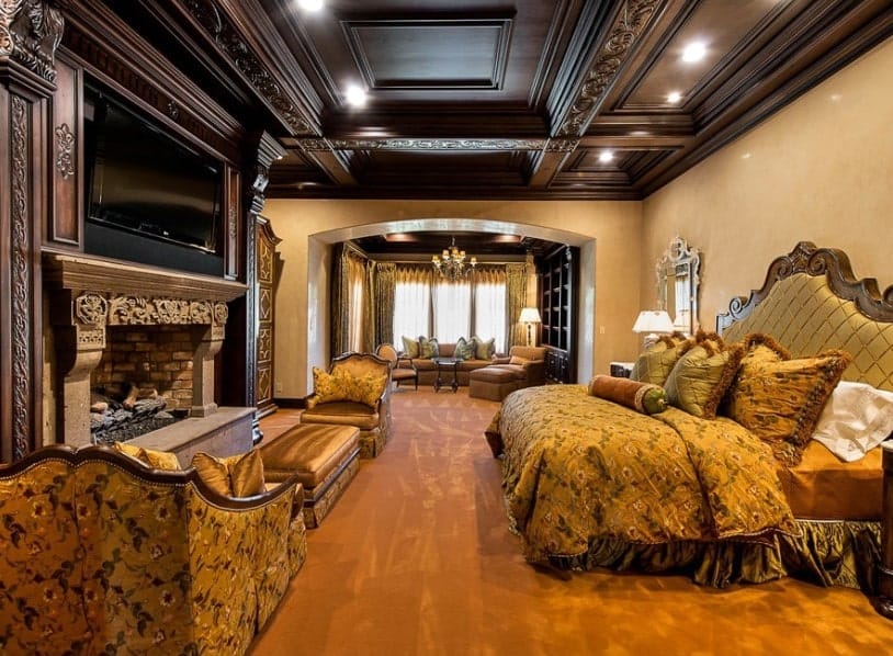 This primary bedroom boasts a stunning decorated ceiling and brown carpet flooring. The room offers a luxurious bed with a large widescreen TV set in front together with a fireplace featuring a sitting area nearby.