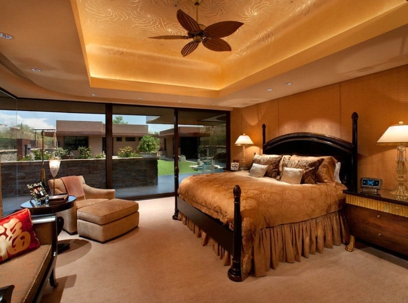 A spacious primary bedroom boasting a stunning decorated tray ceiling, along with brown walls and carpet flooring. The room also offers a large and comfy bed and a sitting space.