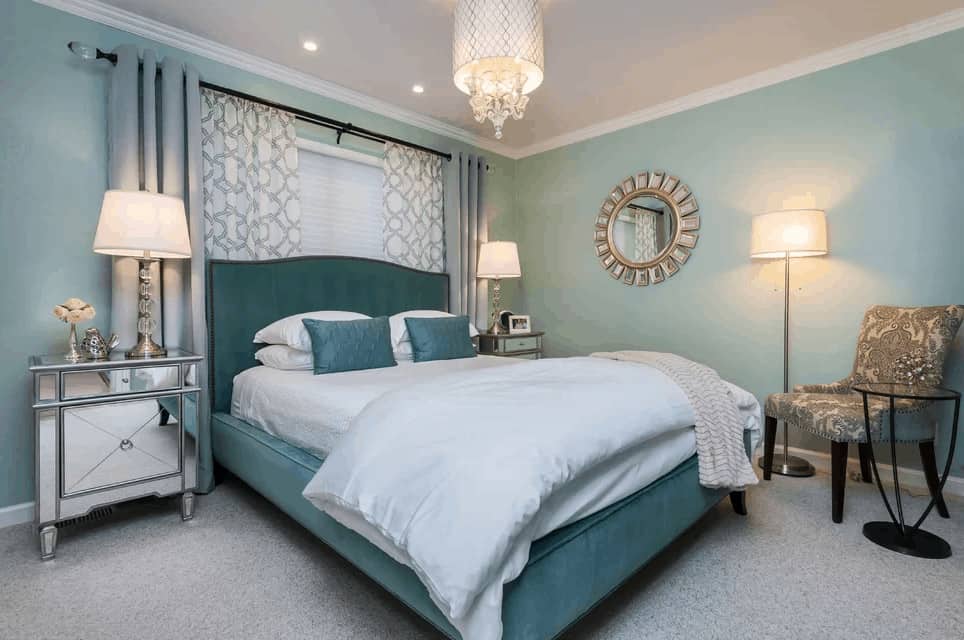 The fabulous primary bedroom features a sunburst mirror and a printed chair facing the green upholstered bed that's illuminated by a lovely drum chandelier. It is flanked by mirrored nightstands and glass table lamps.