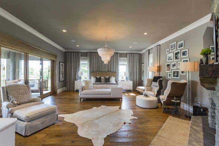 Gray primary bedroom decorated with a photo gallery and a boho chandelier that hung over the cushioned bench. It has a cozy bed and seats along with cowhide and jute rugs that lay on the wide plank flooring.