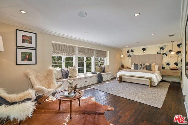 Glass pendant lights illuminate the wooden nightstands flanking a gray tufted bed with a wooden bench on its end. This room boasts a window seat nook and a pair of white faux fur chairs over a cowhide rug.